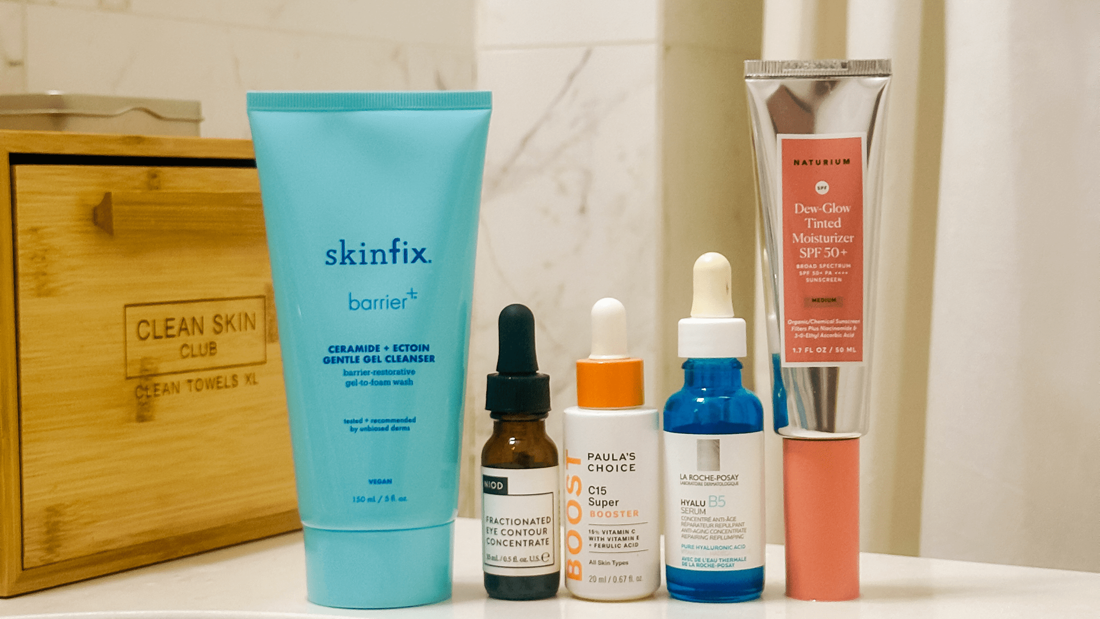 A barrier-supportive skincare routine including Skinfix gentle gel cleanser, La Roche-Posay Hyalu B5 hydrating serum, NIOD eye concentrate, Paula's Choice vitamin C booster, and Naturium SPF 50+ sunscreen arranged on a marble bathroom counter